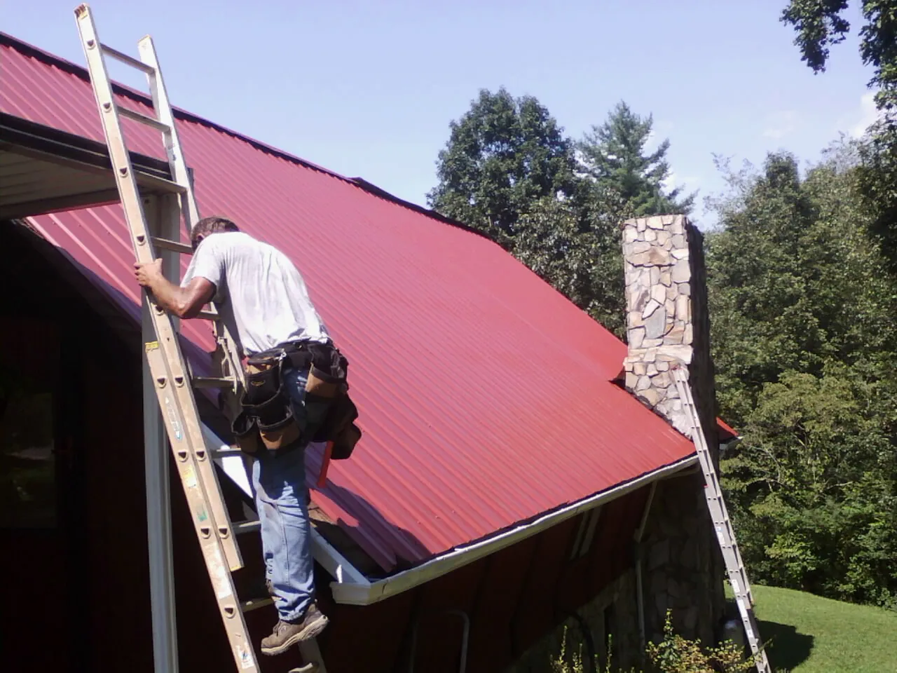 Roofer on a ladder beside a red metal roof and stone chimney — Soffit & Fascia Repair by Roofing Brothers in Wooster